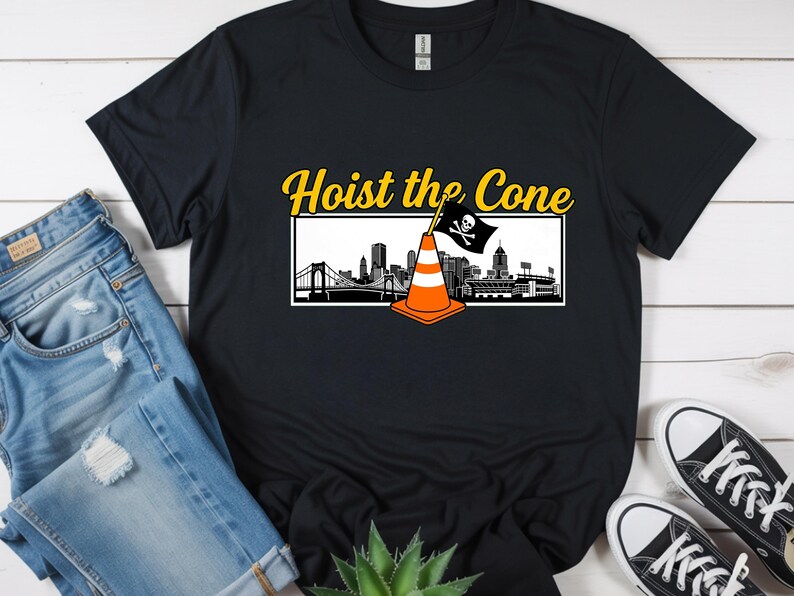 May include: Black t-shirt with the text "Hoist the Cone" in yellow, above a graphic of a traffic cone with a pirate flag and a city skyline. The shirt is paired with blue jeans and black sneakers.