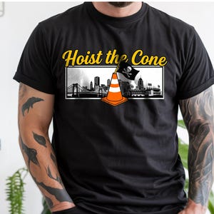 May include: Black t-shirt with the text "Hoist the Cone" in yellow. The design features a traffic cone with a pirate flag and a cityscape. The shirt is a crew neck and short-sleeved.