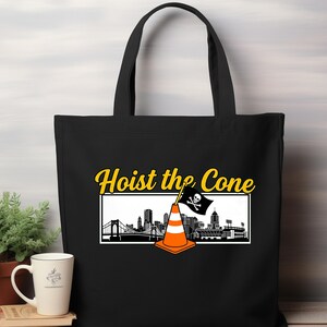 May include: Black tote bag featuring the phrase "Hoist the Cone" in yellow script. The design includes a cityscape silhouette with a traffic cone and a pirate flag. The bag has two black handles.