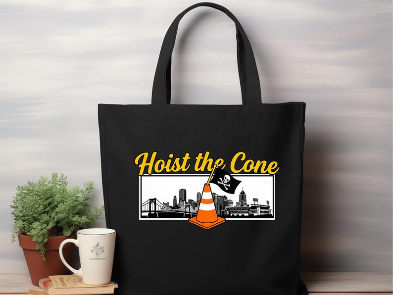 May include: Black tote bag featuring the phrase "Hoist the Cone" in yellow script. The design includes a cityscape silhouette with a traffic cone and a pirate flag. The bag has two black handles.