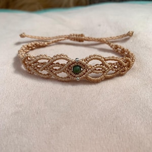 May include: A beige macrame bracelet with an adjustable sliding knot. The bracelet has a central design with a small, round, dark green bead and silver accents. The macrame pattern is intricate and detailed.