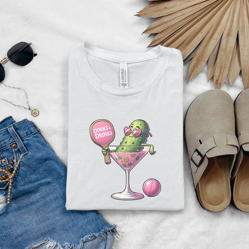 May include: White t-shirt featuring a cartoon pickle wearing sunglasses and holding a pink paddle that says "Dinks & Drinks". The pickle is in a martini glass with a pink ball next to it.