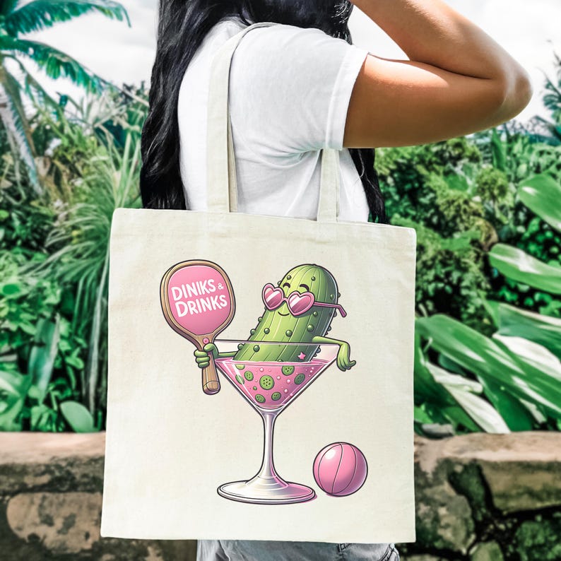May include: A white canvas tote bag featuring a cartoon pickle wearing sunglasses and holding a pink paddle that says "Dinks & Drinks". The pickle is in a pink martini glass with a pink ball next to it.