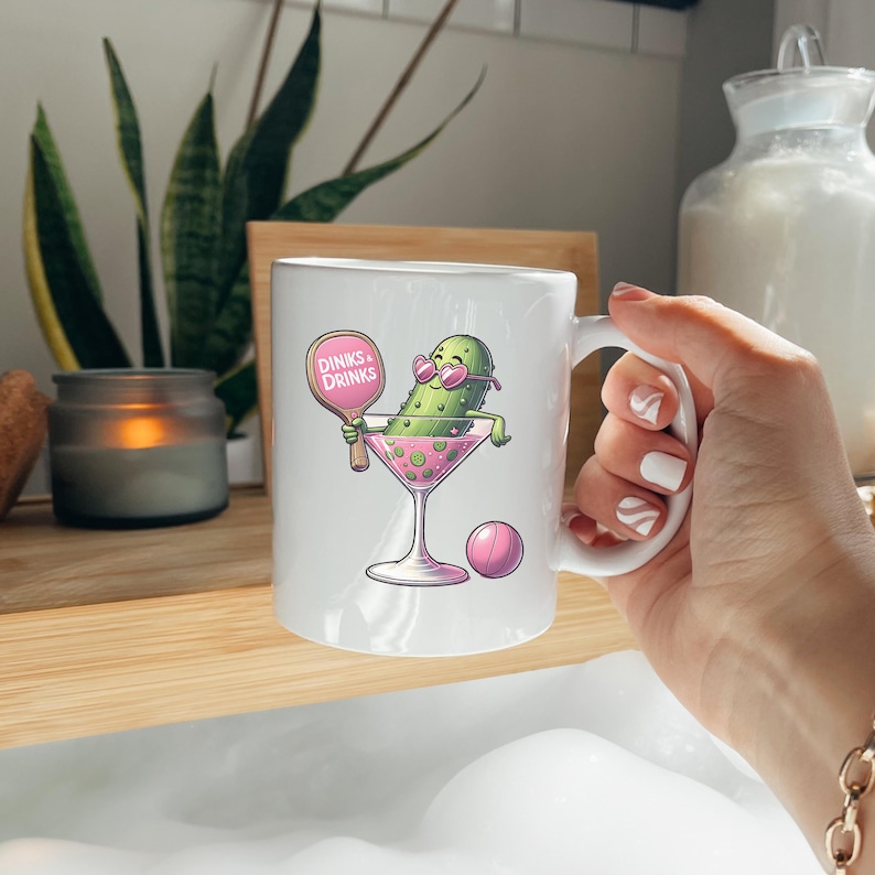 May include: White ceramic mug featuring a green pickle wearing sunglasses and holding a pink paddle with the text "Dinks & Drinks". The pickle is in a pink martini glass with pink olives. There is a pink ball next to the glass.