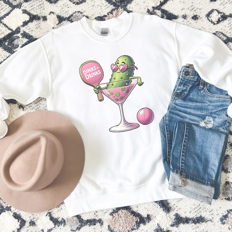 May include: White sweatshirt featuring a pink and green cartoon pickle holding a pink paddle with the text "Dinks & Drinks". The pickle is wearing pink sunglasses and is holding a pink martini glass. There is a pink ball in the background.