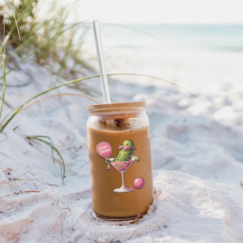 May include: A glass jar with a bamboo lid filled with a brown beverage and a green pickle wearing sunglasses and holding a pink paddle with the text "Drinks & Drinks" on it. The jar has a clear glass straw sticking out of the lid. The jar is sitting on a white sandy beach.