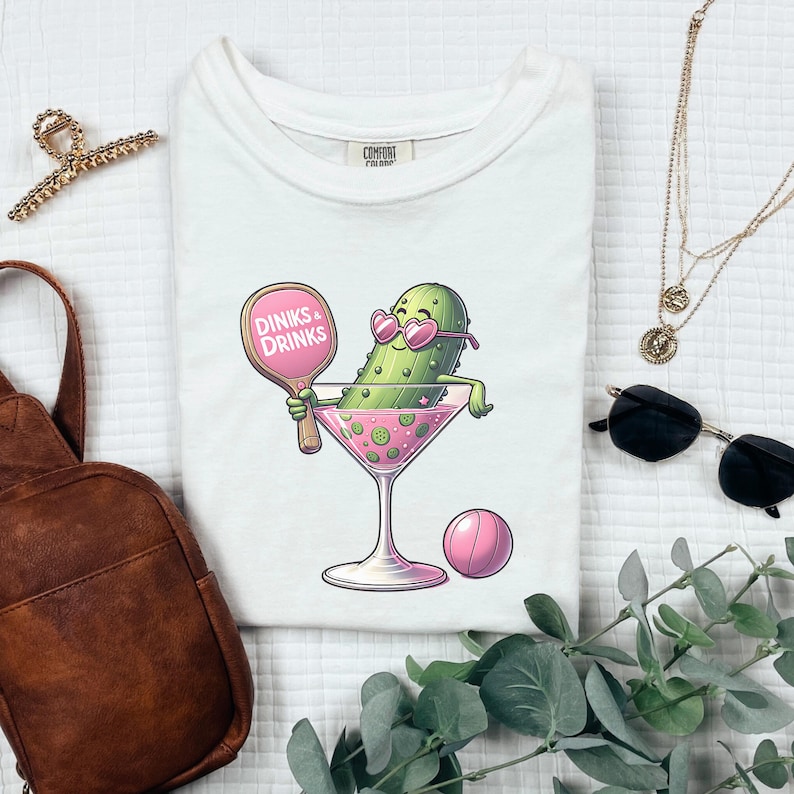 May include: White t-shirt featuring a cartoon pickle wearing sunglasses and holding a pink paddle that says "Dinks & Drinks". The pickle is in a martini glass with a pink rim and a pink ball next to it.