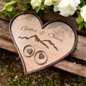 Puede incluir: Una caja de madera para anillos en forma de corazón con un diseño de cordillera y el texto "Gokce & Onur 15.10.2023". Dos anillos están colocados en la caja, uno de oro y otro de plata.