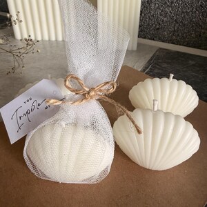 May include: Three off-white shell-shaped candles with vertical ridges. One candle is wrapped in white tulle fabric tied with a brown twine bow and features a white rectangular tag with the black printed text "YOU INSPIRE ME".