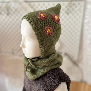 Könnte beinhalten: Olivgrüne Strickhaube mit drei gestickten rosa und gelben Blumen. Die Kapuze hat eine spitze Oberseite und einen integrierten Schal. Das Kleidungsstück wird auf einer Schaufensterpuppe präsentiert, die die handgefertigten Details und die gemütliche Textur zeigt.