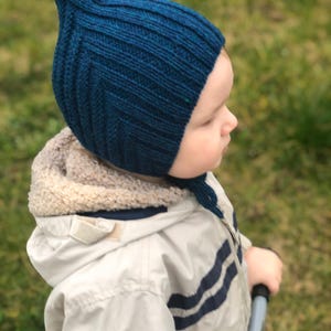 Könnte beinhalten: Eine tealblaue gestrickte Baby-Mütze mit spitzer Spitze. Die Mütze hat eine gerippte Textur und eine Bindung unter dem Kinn. Das Kind trägt eine hellfarbige Jacke mit blauen Streifen.
