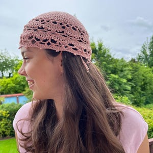 Könnte beinhalten: Eine altrosa gehäkelte Kopfbedeckung mit einem zarten Spitzenmuster und bogenförmigen Kanten. Das Accessoire ist aus feinem Garn gefertigt und weist lose Fadenenden an der Seite auf, die das detaillierte und aufwendige Maschenbild unterstreichen.