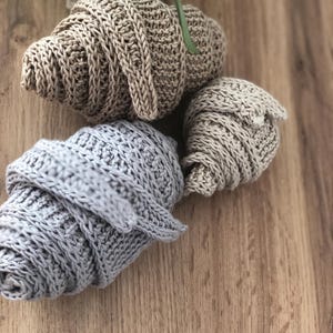 Könnte beinhalten: Drei gehäkelte, croissantförmige Objekte in verschiedenen Größen und Farben. Ein großes beiges Croissant, ein mittelgroßes hellgraues Croissant und ein kleines beiges Croissant sind zusammen angeordnet, jedes mit einem strukturierten Garnmuster.