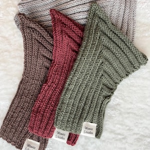 Könnte beinhalten: Nahaufnahme von mehreren Strickartikeln in Braun-, Burgunder- und Olivgrüntönen. Jeder Artikel hat ein Etikett mit der Aufschrift "Mamas Schatze". Die Artikel sind auf einer weißen, strukturierten Oberfläche angeordnet.