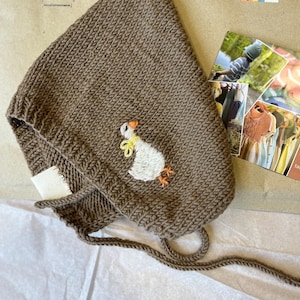 Könnte beinhalten: Eine braune gestrickte Kapuze mit einer weißen Gans, die auf der Vorderseite gestickt ist. Die Kapuze hat einen Kordelzug und ist aus einem weichen, gemütlichen Material gefertigt.