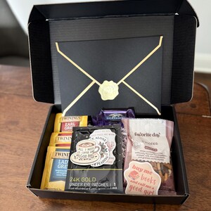 May include: A black gift box with a gold envelope design. The box is open and contains various items, including tea bags, a sticker that says "All I need is coffee and a good book", under eye patches, a bag of s'mores, and a sticker that says "Buy me books and call me a good girl".