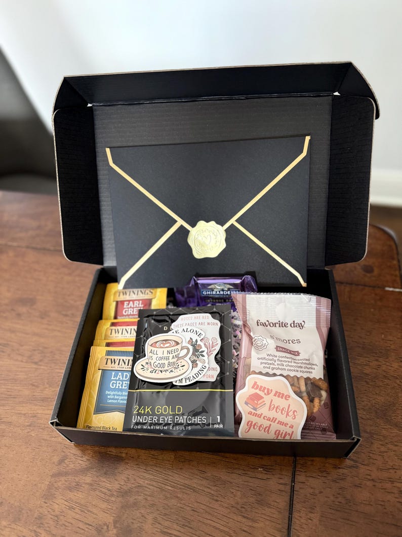 May include: A black gift box with a gold envelope design. The box is open and contains various items, including tea bags, a sticker that says "All I need is coffee and a good book", under eye patches, a bag of s'mores, and a sticker that says "Buy me books and call me a good girl".
