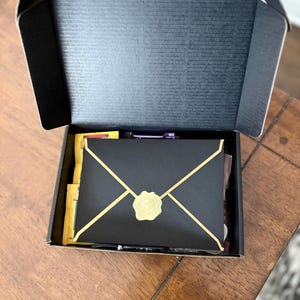 May include: A black box containing a gold envelope. The envelope has a gold wax seal with a design on it.