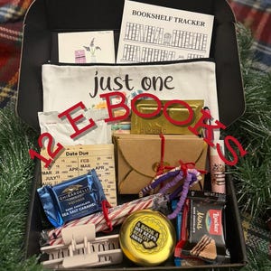 May include: A black box filled with various items, including a white pouch with "just one BOOKS" text, a "Bookshelf Tracker" card, a Ghirardelli chocolate bar, and a gold-colored tin with the text "A BOOK A DAY KEEPS REALITY AWAY."