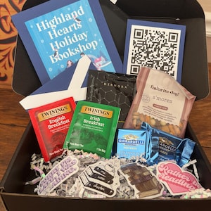 May include: A black gift box filled with various items, including two boxes of Twinings tea, a bag of s'mores snack mix, two Ghirardelli chocolate squares, and several book-themed stickers. The box also includes a blue card with a QR code and a blue card with the text "Highland Hearts Holiday Bookshop".