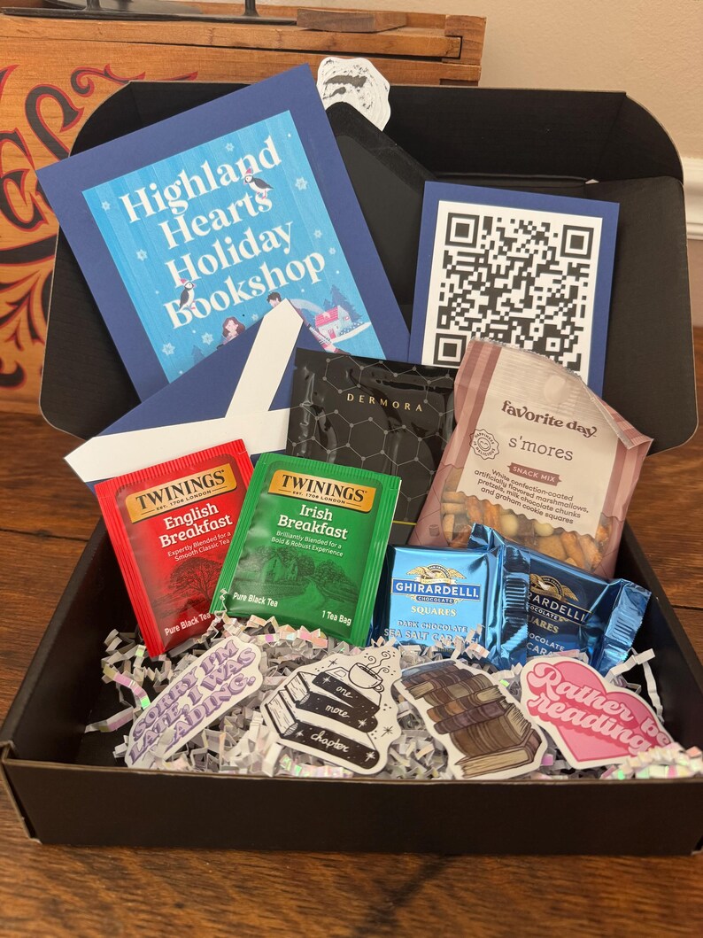 May include: A black gift box filled with various items, including two boxes of Twinings tea, a bag of s'mores snack mix, two Ghirardelli chocolate squares, and several book-themed stickers. The box also includes a blue card with a QR code and a blue card with the text "Highland Hearts Holiday Bookshop".