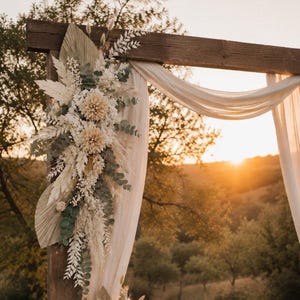 May include: A wooden wedding arch decorated with a large floral arrangement of dried palm leaves, pampas grass, white flowers, and green eucalyptus foliage, featuring draped sheer white fabric hanging from the top horizontal beam.
