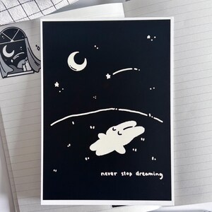 May include: A black and white art print featuring a whimsical illustration of a creature floating in a starry night sky with a crescent moon. The text "never stop dreaming" is at the bottom. The print is on a white background.