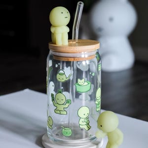 Op de afbeelding: Een heldere glazen beker in de vorm van een blikje met een bamboe deksel en een glazen rietje. De beker is versierd met groene cartoonfiguren. Twee groene figuren zijn op het deksel en de basis geplaatst. De achtergrond bevat een witte konijnenlamp.