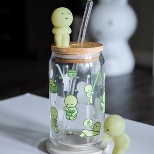 Op de afbeelding: Een doorzichtige glazen beker met een bamboe deksel en een doorzichtig glazen rietje. De beker is versierd met groene cartoonfiguren. Twee kleine, lichtgroene beeldjes zijn op het deksel en naast de beker geplaatst.