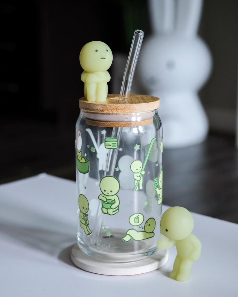 Op de afbeelding: Een doorzichtige glazen beker met een bamboe deksel en een doorzichtig glazen rietje. De beker is versierd met groene cartoonfiguren. Twee kleine, lichtgroene beeldjes zijn op het deksel en naast de beker geplaatst.