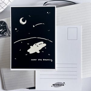 May include: A black and white postcard featuring a whimsical illustration of a creature floating in a starry night sky with a crescent moon. The text "never stop dreaming" is written below the illustration. The back of the postcard is white with a stamp box and the artist's logo.