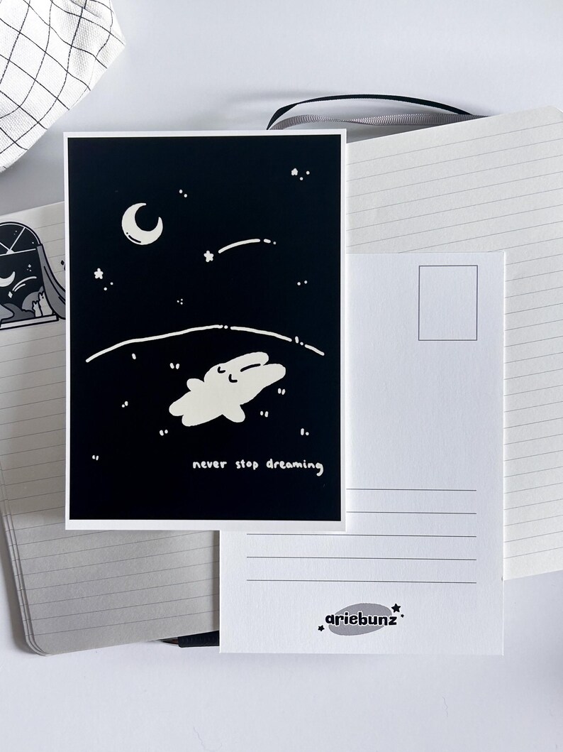 May include: A black and white postcard featuring a whimsical illustration of a creature floating in a starry night sky with a crescent moon. The text "never stop dreaming" is written below the illustration. The back of the postcard is white with a stamp box and the artist's logo.