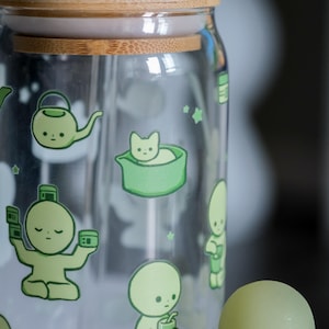 Op de afbeelding: Een heldere glazen pot met een bamboe deksel en een rietje. De pot is versierd met groene cartoon illustraties van een theepot, een kat in een kom en andere figuren. De pot is ontworpen voor koude dranken.