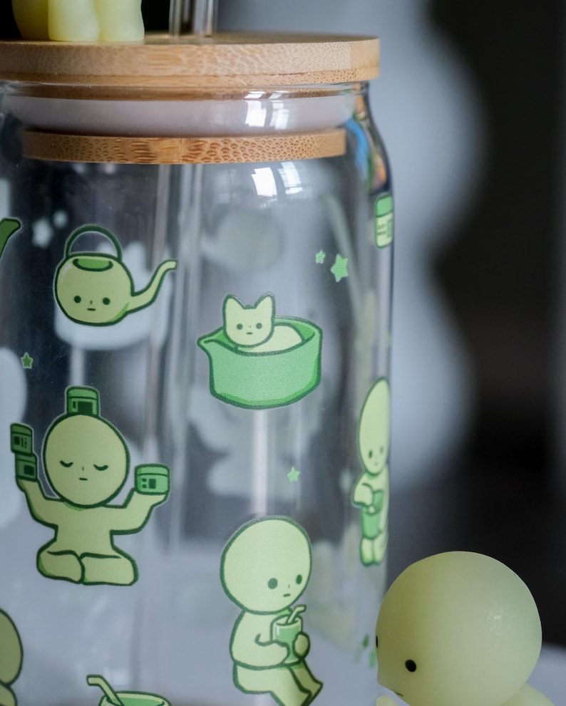 Op de afbeelding: Een heldere glazen pot met een bamboe deksel en een rietje. De pot is versierd met groene cartoon illustraties van een theepot, een kat in een kom en andere figuren. De pot is ontworpen voor koude dranken.