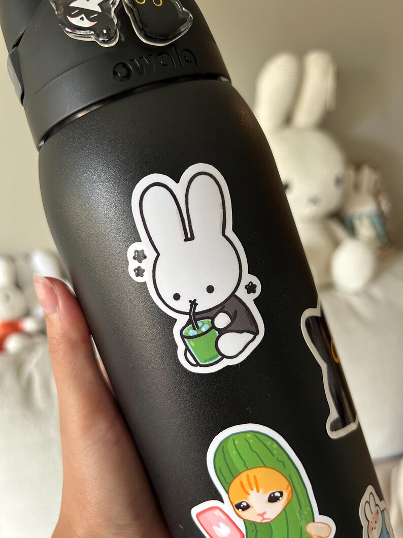 May include: Black Owala water bottle decorated with cartoon stickers. A white bunny sticker drinks from a green cup. Other stickers include a black cat and a cat in a cucumber costume. The bottle is held by a hand.