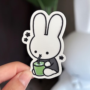 May include: A white bunny sticker with a black outline, drinking from a green cup. The bunny is wearing a black shirt and has three black stars next to it. The sticker is held by a hand.