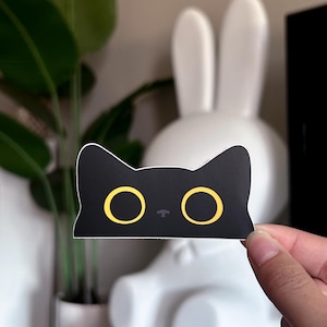 May include: A black cat sticker with yellow eyes and a white border. The sticker is held up in front of a white bunny statue and a potted plant. The cat sticker is a cute and simple design.