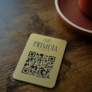 May include: A gold-coloured card with the word "PRÍMULA" and "CARTA" printed in black. The card features a QR code and is placed on a wooden surface. A red saucer and cup are visible in the background.