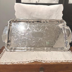 May include: A rectangular silver-toned serving tray featuring an ornate, embossed floral and scrollwork pattern across its surface. The tray has a raised, scalloped edge with integrated handles on both sides and a polished, reflective finish.
