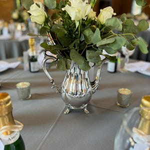 May include: A silver teapot with a floral arrangement of white roses and green leaves. The teapot is sitting on a gray tablecloth with two bottles of champagne and two candle holders.