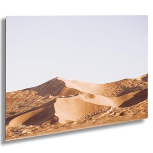 May include: A landscape photograph of golden sand dunes under a clear, pale sky. The dunes feature sharp ridges and deep shadows, creating a textured surface with undulating curves. A small, faint signature is visible in the bottom left corner.