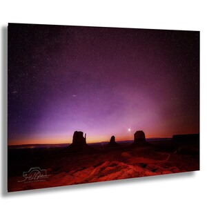 May include: A landscape photograph of Monument Valley at night. The image features a dark purple and black starry sky above the red rock formations. The foreground is a reddish-brown desert landscape. The photograph is signed in the lower left corner.