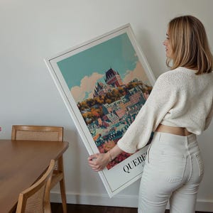 May include: A white framed print of a colorful illustration of a city skyline with the word "QUEBEC" at the bottom. A person is holding the print in front of a white wall.