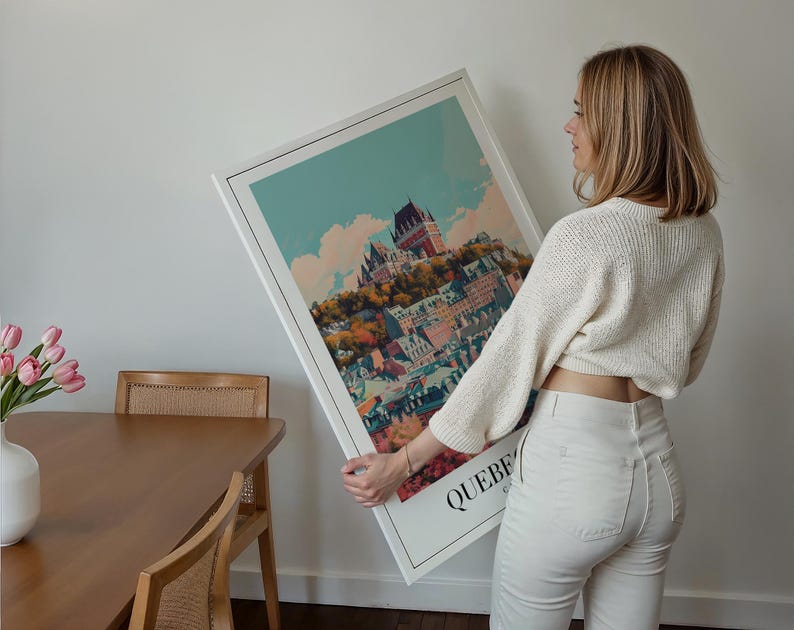 May include: A white framed print of a colorful illustration of a city skyline with the word "QUEBEC" at the bottom. A person is holding the print in front of a white wall.