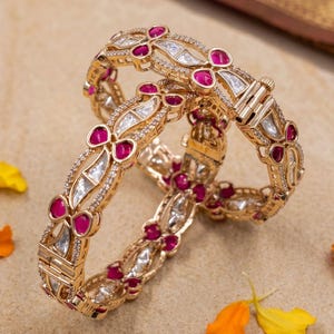 May include: Two gold-colored bangles with intricate designs. Each bangle features alternating ruby-colored and clear, sparkling stones. The bangles are adorned with small, clear stones and are displayed on a neutral surface with scattered flower petals.
