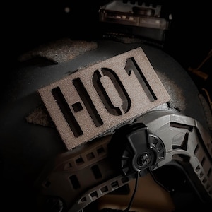 Könnte beinhalten: Nahaufnahme eines beigen taktischen Helms mit einem braunen Patch mit der Aufschrift "H01", der an der Seite befestigt ist.