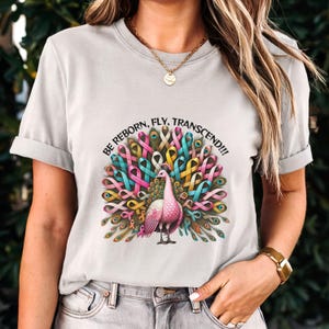 Puede incluir: Una camiseta blanca con un diseño de pavo real hecho de cintas de colores. El pavo real es rosa y tiene una cola verde y dorada. El texto "BE REBORN, FLY, TRANSCEND!!!" está impreso sobre el pavo real.