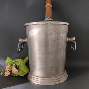 May include: Silver metal champagne bucket with two black metal handles and a bottle of champagne inside.