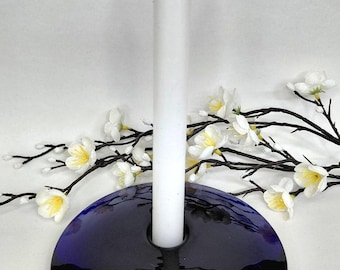 Peut inclure: Une bougie blanche dans un bougeoir en verre bleu foncé et noir. Le bougeoir est rond avec un trou central pour la bougie. Des fleurs et des branches blanches sont disposées autour de la bougie et du bougeoir.