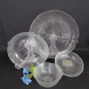 May include: A collection of four clear glass serving dishes, each shaped like a leaf with textured veining. The set includes a large platter, two bowls, and a smaller bowl. A small bouquet of blue flowers is placed near the dishes.